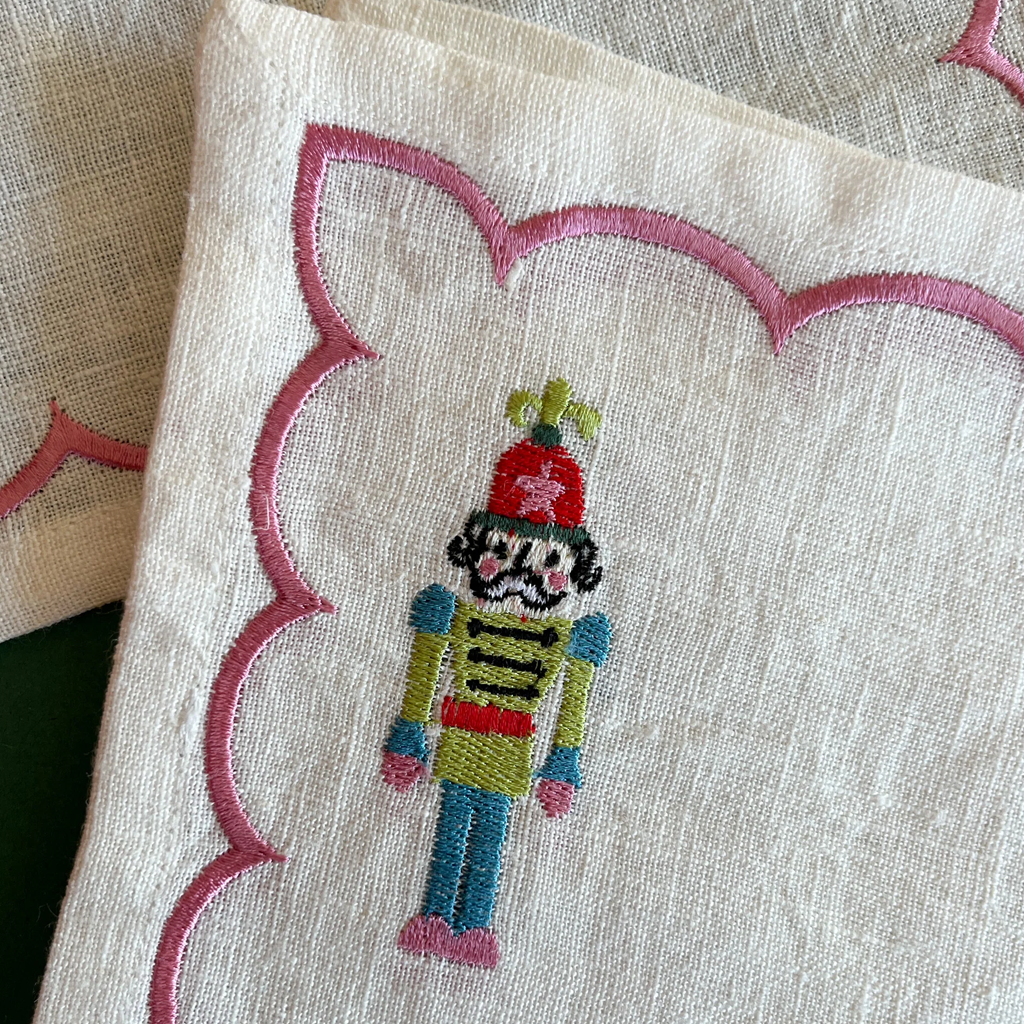 Close-up of a cream-colored linen napkin with a pink scalloped border and an embroidered nutcracker figure in the center.