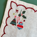 A close-up of a cream-colored linen napkin with a scalloped red border and embroidered Christmas ornaments in the center, set against a dark green background.