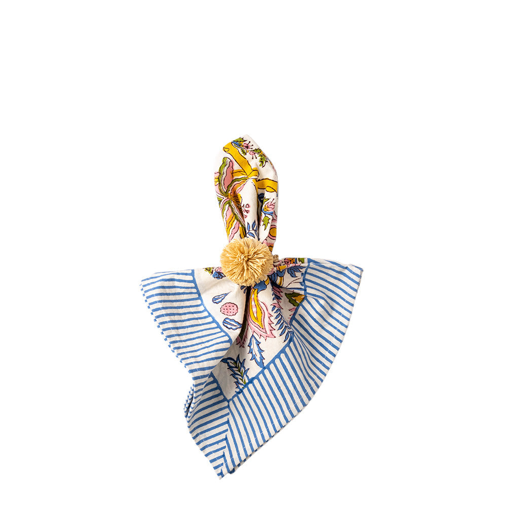 A folded napkin with a blue and white striped pattern on one side and a multicolor print on the other, tied in the center with a yellow pom-pom napkin ring, shown on a plain white background.