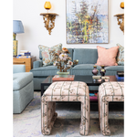 A styled living room scene featuring two upholstered ottomans with a beige floral pattern in the foreground, a blue-gray sofa with patterned and pink pillows in the background, and decorative wall sconces and framed art.