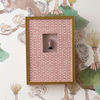 A square framed wall art piece with a gold frame and pink patterned backing, featuring a small central inset showing a miniature bottle. The piece is displayed against a decorative botanical wallpaper.