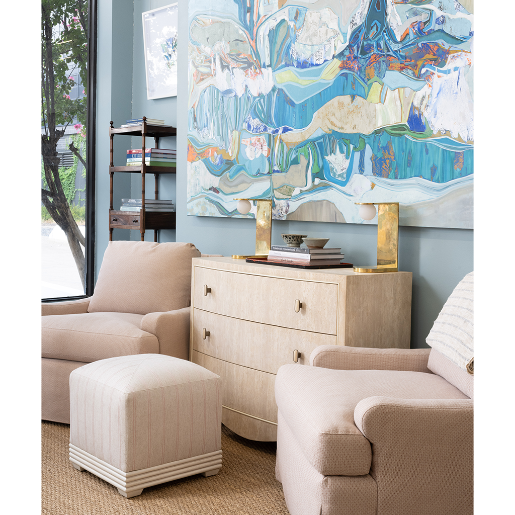 A styled interior scene with blush beige upholstered armchairs, a matching ottoman, a light wood chest of drawers with gold-tone hardware, and a large abstract wall artwork above. No people or animals are present, but this is still not a clean isolated product shot because it shows furniture arranged in a real room setting, so subject_type is Lifestyle.