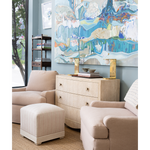 A styled interior scene with blush beige upholstered armchairs, a matching ottoman, a light wood chest of drawers with gold-tone hardware, and a large abstract wall artwork above. No people or animals are present, but this is still not a clean isolated product shot because it shows furniture arranged in a real room setting, so subject_type is Lifestyle.