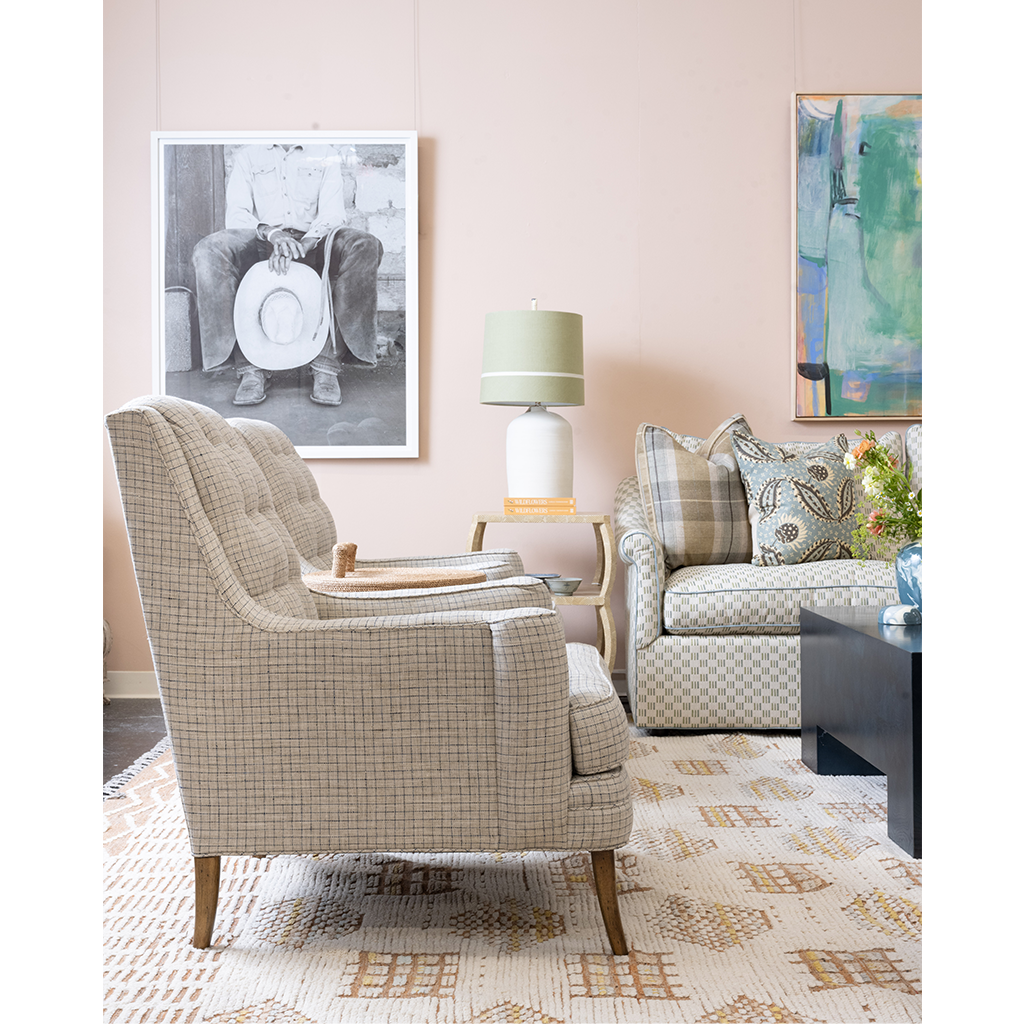 A styled living room scene featuring a beige and white checkered tufted armchair in the foreground, a matching patterned sofa in the background, a patterned area rug, a table lamp, and framed wall art against a soft blush pink wall.