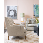 A styled living room scene featuring a beige and white checkered tufted armchair in the foreground, a matching patterned sofa in the background, a patterned area rug, a table lamp, and framed wall art against a soft blush pink wall.