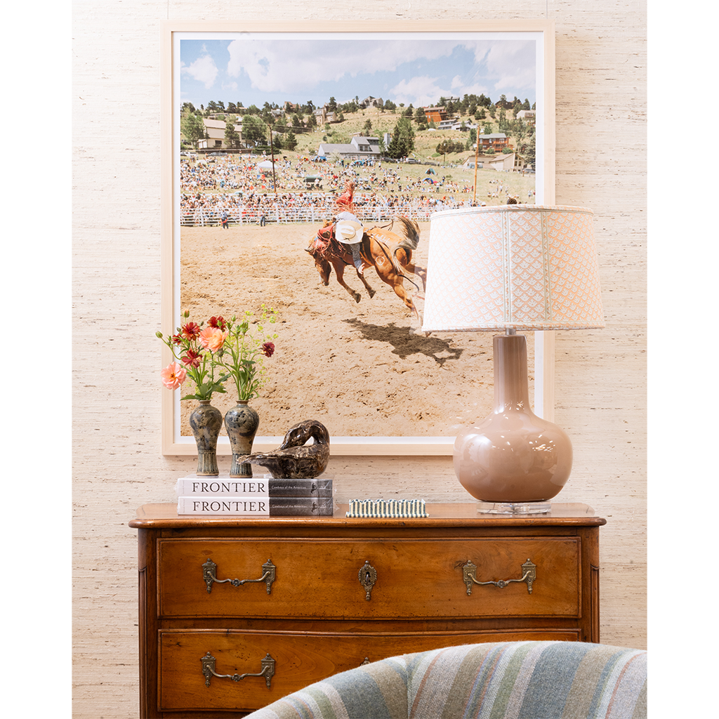 A large framed art print depicting a horse race scene, styled in a home decor setting above a wooden dresser with a lamp, books, and vases.