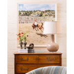 A large framed art print depicting a horse race scene, styled in a home decor setting above a wooden dresser with a lamp, books, and vases.