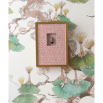 A framed decorative wall piece with a pink patterned insert and a small central figure, mounted against wallpaper featuring green and beige botanical branches.