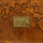 An antique wooden box lid featuring a rich, swirling burl wood pattern in warm brown and orange tones, with a central rectangular metal inset showing a vintage illustration and the text 'true to you'.