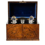 An antique wooden presentation box with a richly figured burl wood exterior, an open dark blue velvet-lined lid, and three clear crystal decant cups resting inside on a black base.