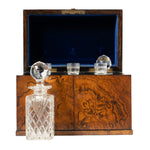 An open burl wood liquor box with a blue velvet-lined interior is displayed alongside a cut crystal decanter with a faceted stopper. Inside the box are fitted holders for glassware, highlighting its elegant design for storing and serving spirits.