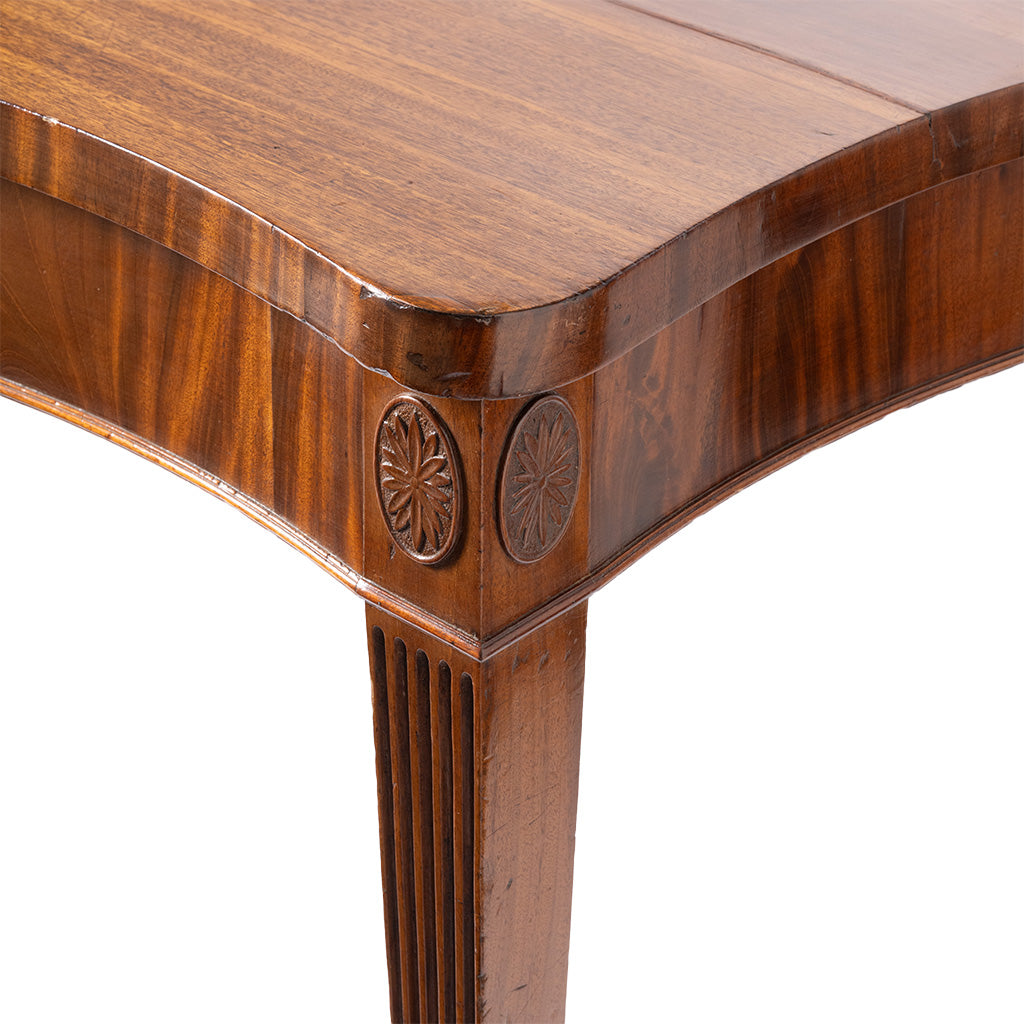 Close-up of a polished wooden table corner with a fluted leg and carved circular medallions on the apron, shown against a white background.