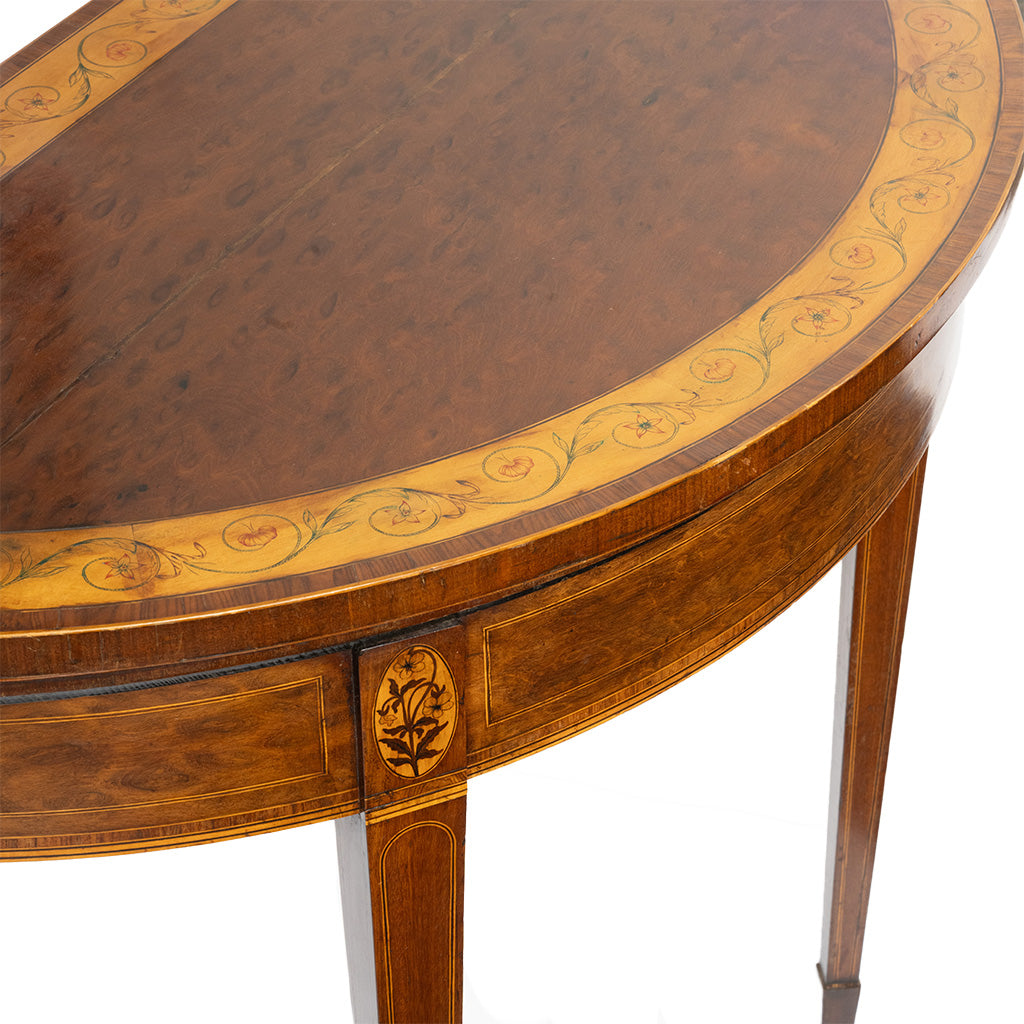 Close-up of an antique wooden demi-lune table showcasing its richly figured wood grain and an intricate inlaid border of scrolling vines and floral motifs around the semicircular top. Delicate inlay on the apron and tapered legs highlights the table's fine craftsmanship.