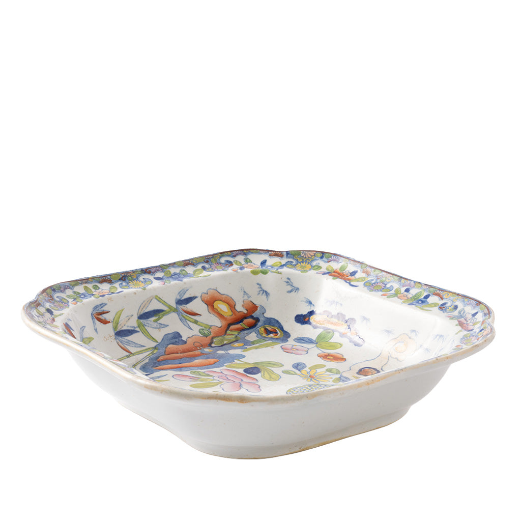 A square ceramic serving bowl with gently flared sides features a colorful hand-painted floral and botanical design in blue, green, orange, pink, and yellow on a white background. An intricate floral border decorates the rim, giving the piece a vibrant, antique-inspired appearance.