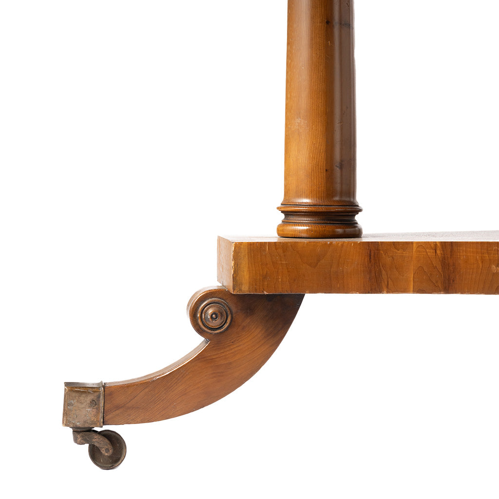 Close-up of the lower corner of a wooden table leg assembly with a turned column, apron, curved bracket, and caster wheel on a white background.