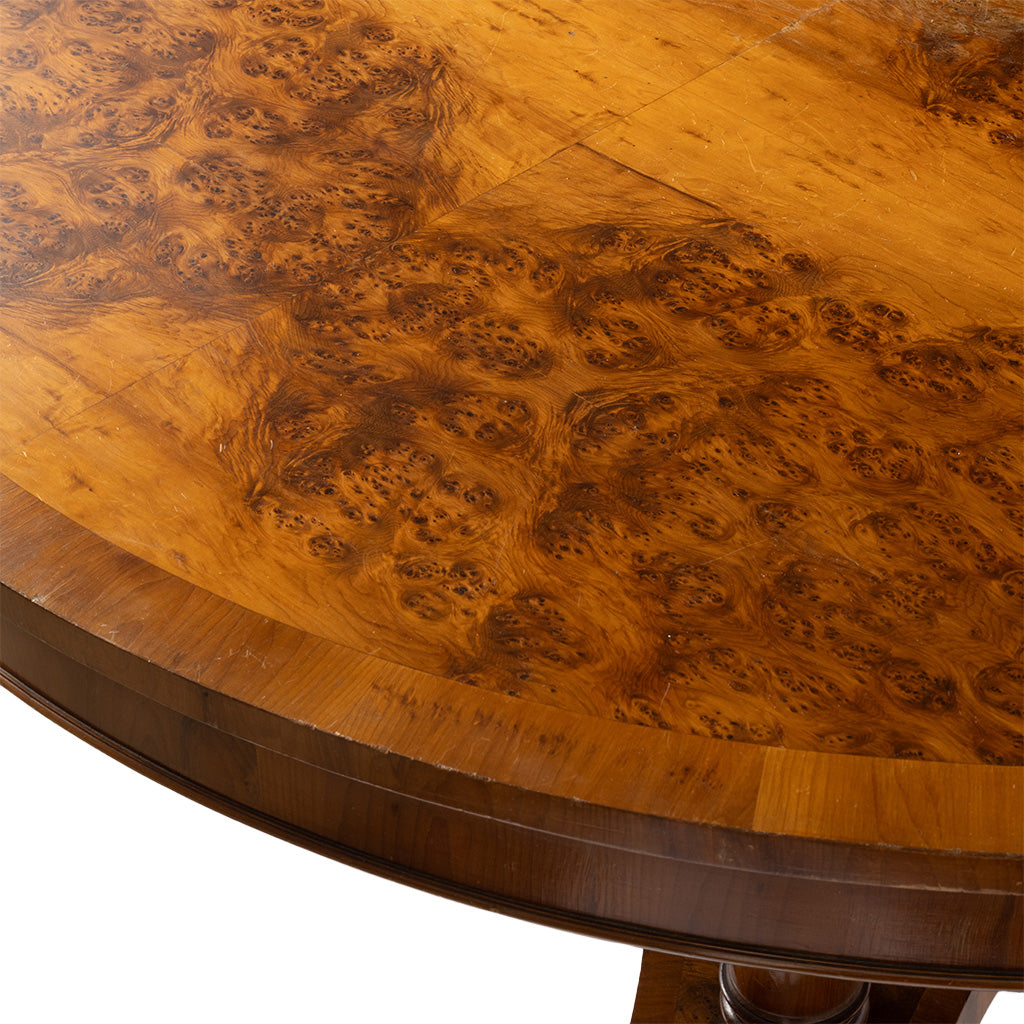 Close-up of a round wooden table top with rich burl wood grain in warm golden-brown tones, showing the polished rim and part of the pedestal base against a white background.