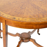 Close-up of a round wooden table with a richly grained flamed wood top, decorative inlay around the rim, and a central pedestal base on casters.