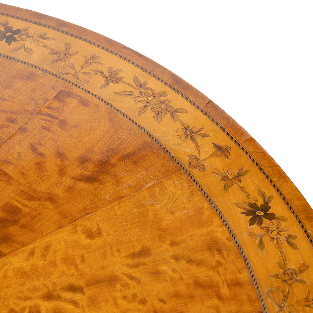 Close-up of a round wooden table showing a warm honey-brown flamed wood surface with an inlaid floral border.