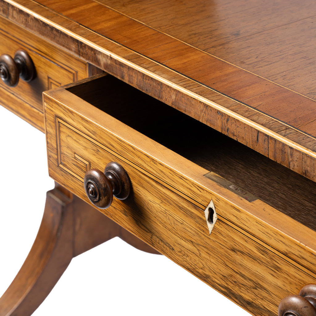 Close-up of a wooden writing desk with an open drawer, showcasing a warm medium-brown wood grain finish, decorative inlay trim, and dark metal drawer knobs on a white background.