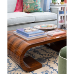 A curved wooden coffee table with a glossy dark brown finish, styled in a living room setting with books, a decorative plate, and a light blue sofa in the background.