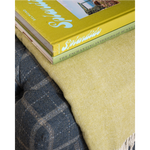 A close-up of a textured herringbone throw blanket in a muted olive-yellow color, styled on a dark gray plaid sofa with two books stacked on top.