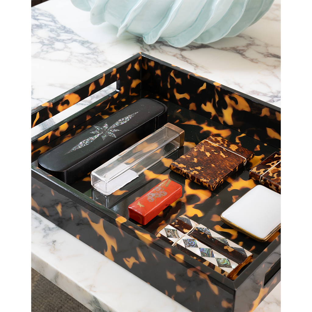 A rectangular tray with a glossy black and orange tortoiseshell pattern, styled with various small accessories and cosmetics on a white marble surface.