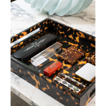 A rectangular tray with a glossy black and orange tortoiseshell pattern, styled with various small accessories and cosmetics on a white marble surface.