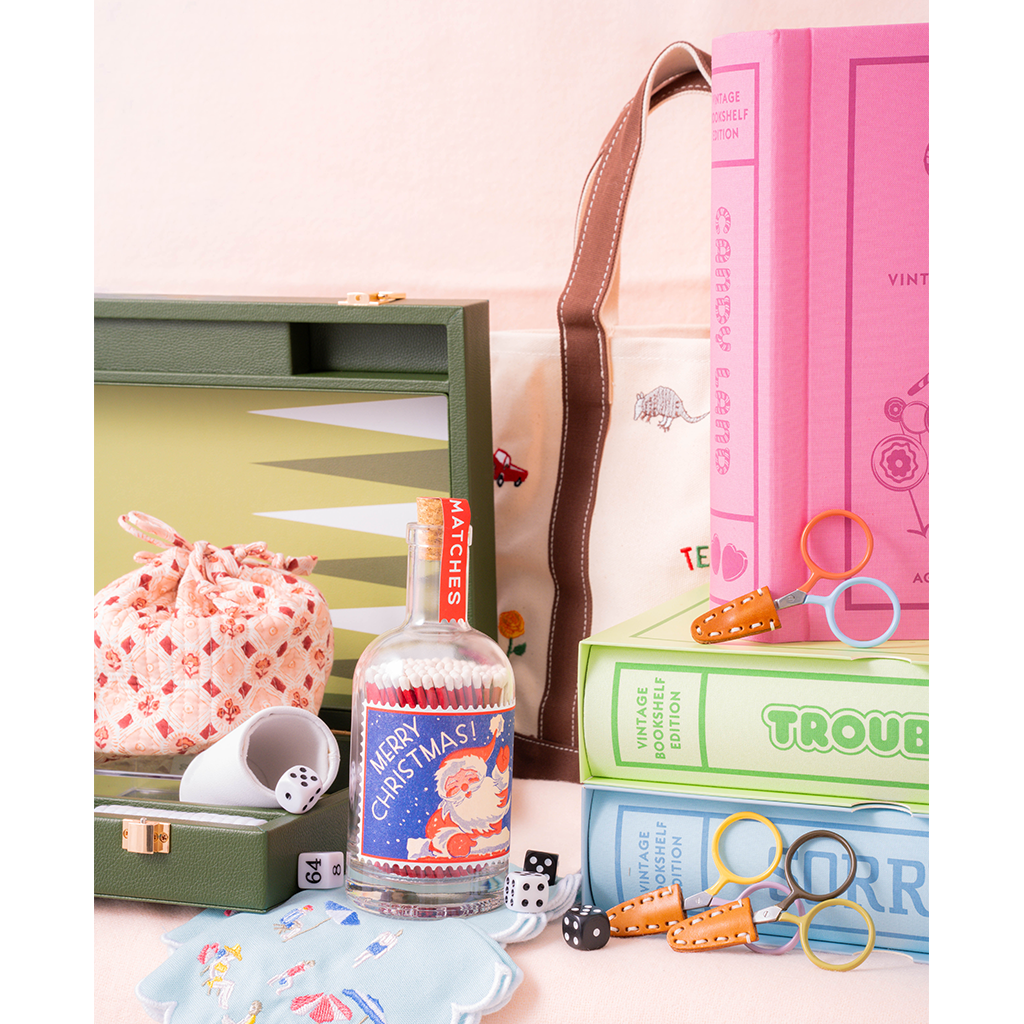 A styled tabletop arrangement of vintage-inspired game accessories and books: an olive green backgammon board set, pink and blue game books, a small pink patterned drawstring bag, a clear bottle with a red-and-blue Christmas label reading 'Merry Christmas,' orange-handled scissors, dice, and a light blue patterned dish.