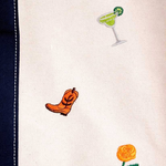 Close-up of a white embroidered fabric with small stitched motifs including a green margarita glass, an orange cowboy boot, and part of an orange flower, next to a dark navy stitched border.