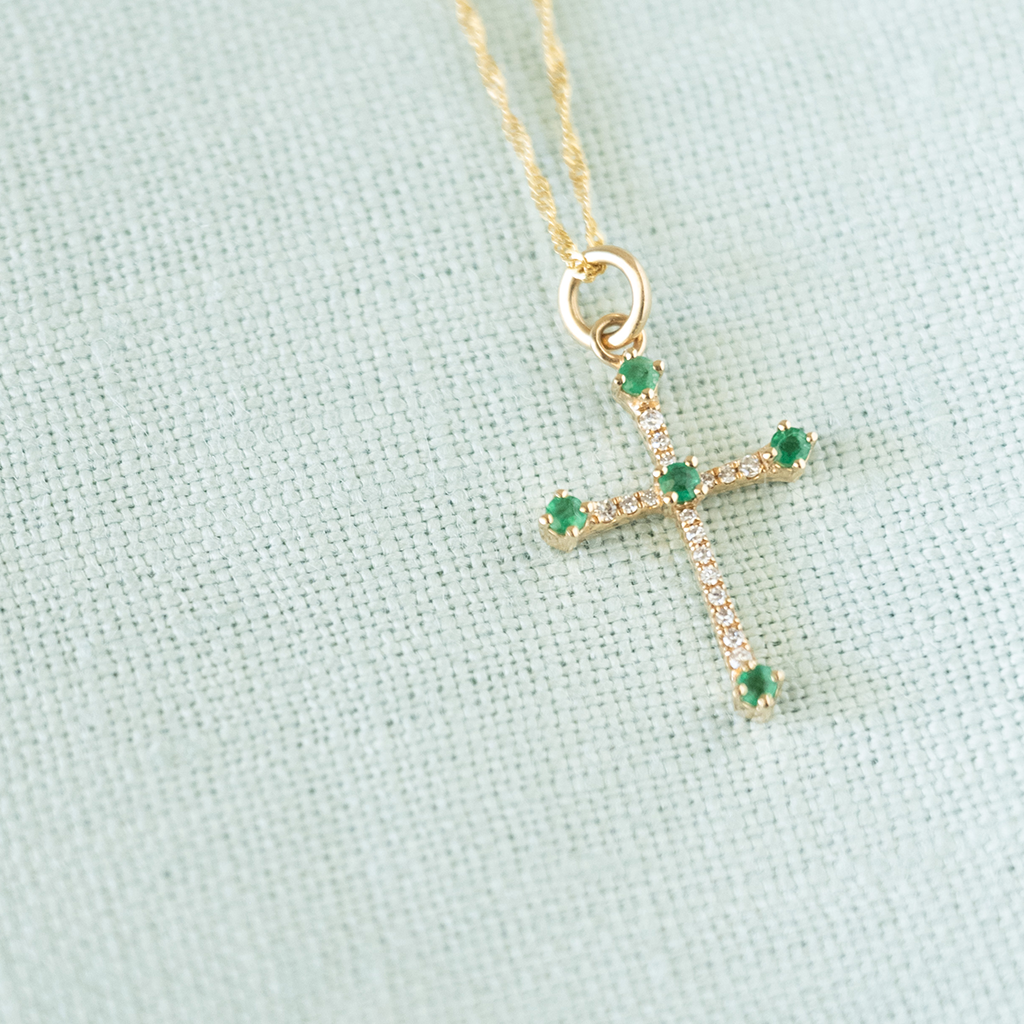A gold cross pendant necklace with small green gemstones and clear stones, displayed on a textured light blue fabric background.