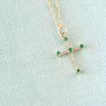 A gold cross pendant necklace with small green gemstones and clear stones, displayed on a textured light blue fabric background.