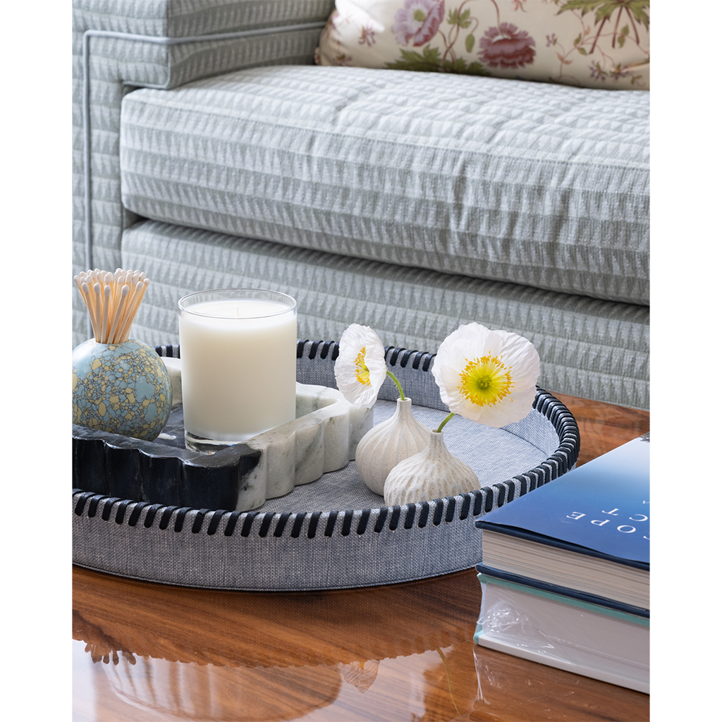 A round gray fabric-covered tray with black braided trim sits on a wooden table in a living room setting, styled with a candle, marble slice, small vase, flowers, and books. A striped sofa is visible in the background.
