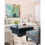 A styled living room scene with a beige sectional sofa, patterned throw pillows, a dark wood coffee table, a blue plaid accent chair, a rug, and a large abstract painting on the wall.