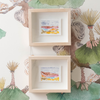 Two light wood shadow-box frames with white mats, each containing a small colorful landscape watercolor, displayed on a white wallpaper with botanical illustrations.
