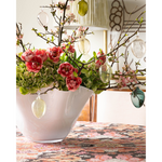 A set of light glass Easter eggs in various shades of yellow and green, placed on a table with a floral arrangement in the background.