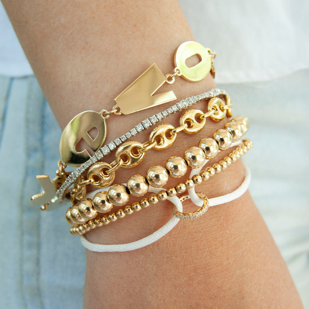 Stack of five gold bracelets featuring varied designs including linked chains, round beads, and a toggle clasp on a wrist.