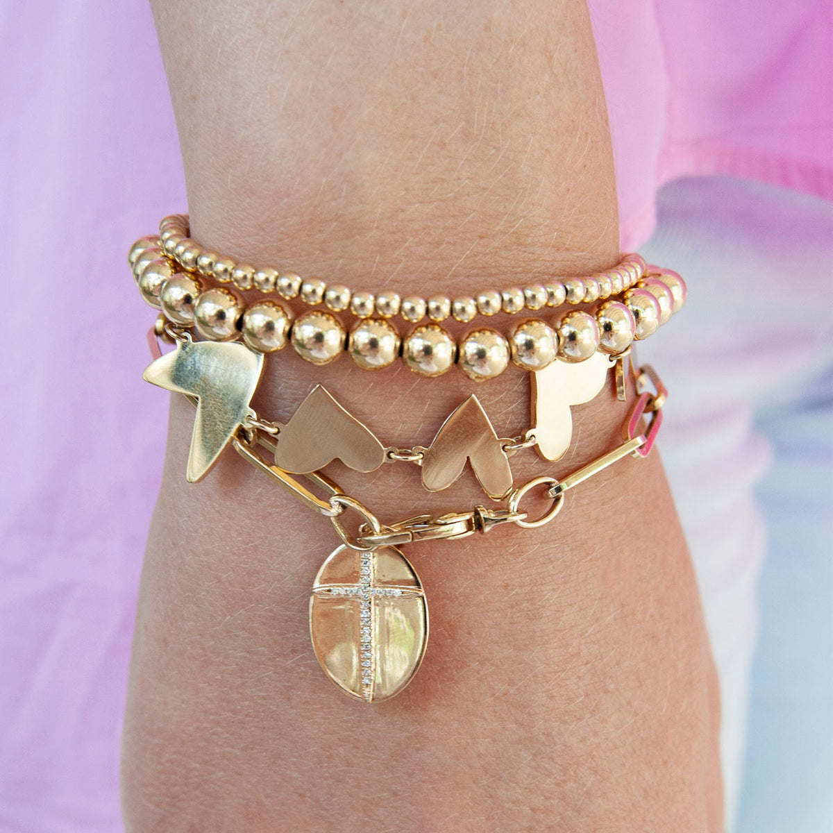 Gold large paperclip chain bracelet with round charm, stacked with beaded and heart bracelets.