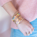 Gold "I Love You" bracelet with delicate chain and subtle engraved detail on wrist alongside colorful beaded and gold bangles.