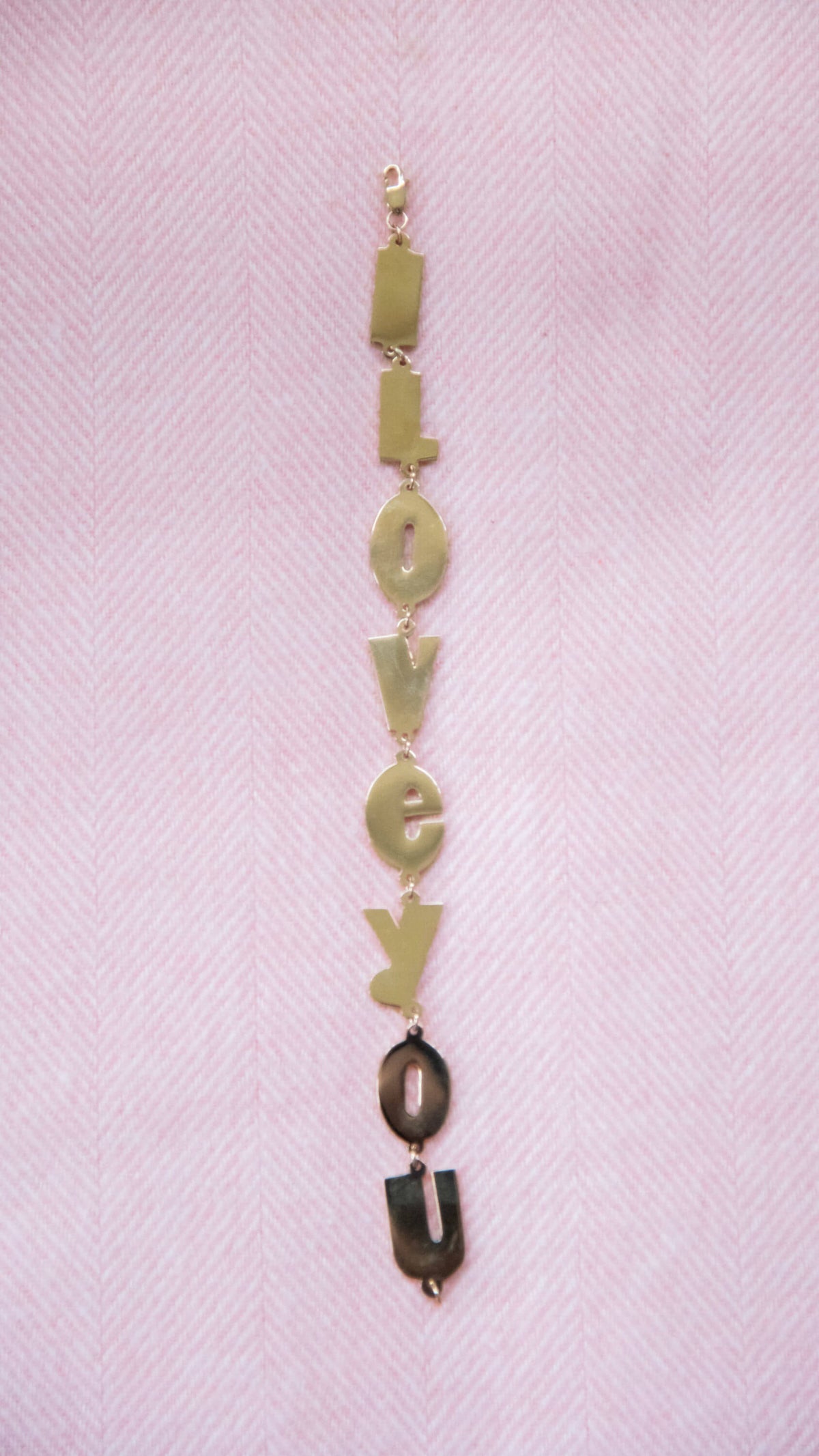 Gold "I Love You" letter charm bracelet on soft pink background.