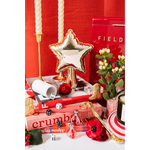 A styled tabletop arrangement featuring a metallic silver star-shaped balloon on a gold stand, red and pink books, white pillar candles, small perfume bottles, dice, flowers, and a red-and-white striped plate against a red backdrop.