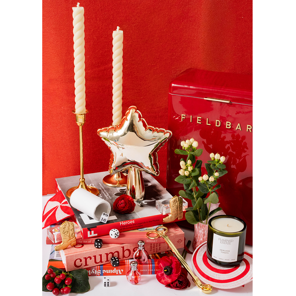 A styled holiday gift arrangement featuring a red Fieldbar tin, two twisted white taper candles in gold holders, a metallic gold star-shaped candle holder, a white candle, books, a small potted plant, a candle in a striped tin, and red poppies against a red background.
