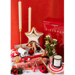 A styled holiday gift arrangement featuring a red Fieldbar tin, two twisted white taper candles in gold holders, a metallic gold star-shaped candle holder, a white candle, books, a small potted plant, a candle in a striped tin, and red poppies against a red background.