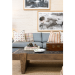 A cozy living room featuring a blue upholstered sofa layered with floral and striped pillows beneath framed black-and-white artwork. A substantial wood cocktail table with a dark stone top is styled with stacked books, decorative objects, and a vintage wooden box.