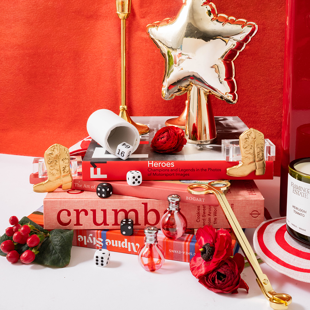 A styled tabletop arrangement featuring a red candle jar labeled 'Pine Ridge Escape' on a striped coaster, surrounded by decorative props including red roses, dice, miniature cowboy boots, books, and a gold star-shaped object against a red backdrop.