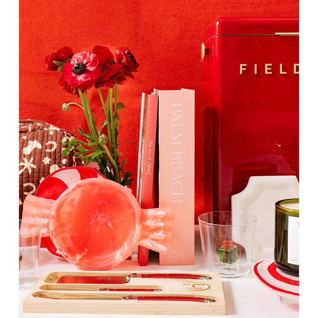 A styled tabletop scene with a red FIELD refrigerator, red flowers, a pink book, a pink glass bowl, a glass, and a wooden tray holding gold-toned utensils with red handles, set against a red wall.