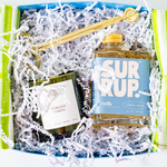 A gift box filled with white shredded paper containing a blue-labeled SURUP vanilla simple syrup bottle, a dark green candle jar labeled Hannah's Estate Heirloom Tomato, and a gold spoon resting across the top.