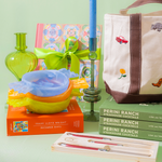 A styled gift set featuring a canvas tote bag with brown straps and colorful embroidered motifs, a green glass bottle, a blue candle, a colorful patterned box tied with a green ribbon, a yellow and orange bowl, and several Perini Ranch Steakhouse cocktail books.