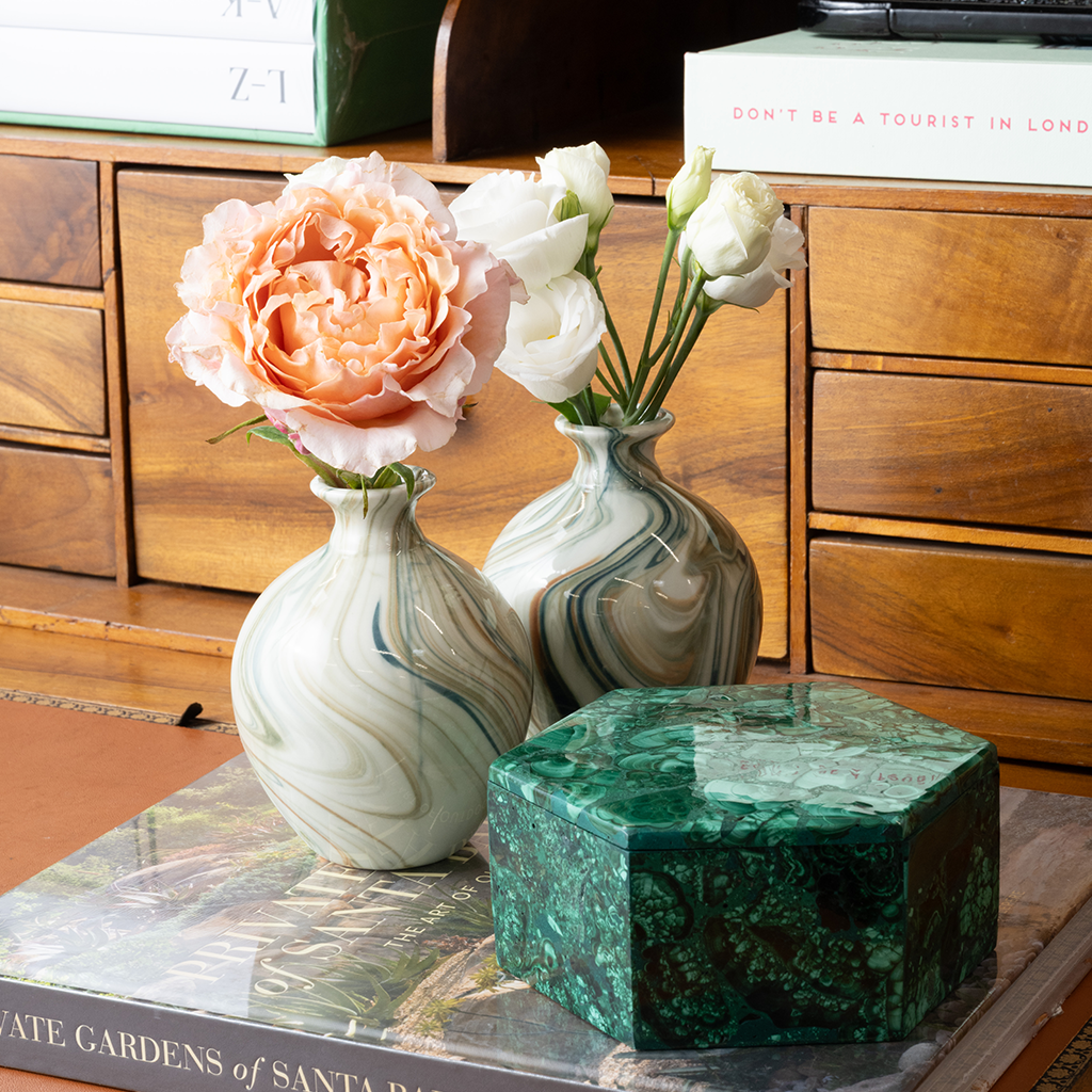 A styled home decor scene featuring two marbled ceramic vases with flowers, set against a wooden dresser background with books.