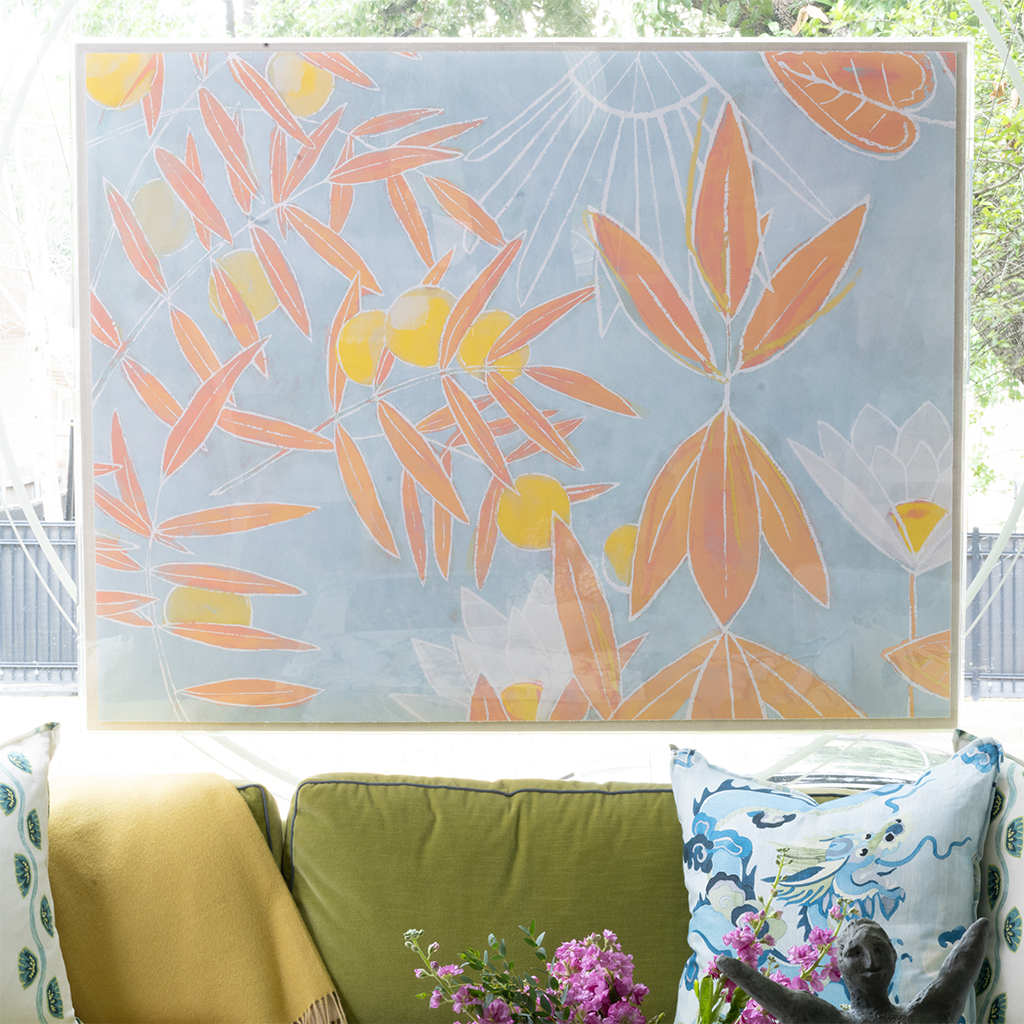 A large framed wall art print featuring a light blue background with orange and yellow botanical leaves and sun-like line details, displayed above a green sofa with decorative pillows.