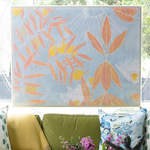A large framed wall art print featuring a light blue background with orange and yellow botanical leaves and sun-like line details, displayed above a green sofa with decorative pillows.
